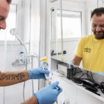 Operatore domiciliare BeeCare con guanti azzurri prepara uno spazzolino con dentifricio davanti a uno specchio in bagno, pronto ad assistere un paziente.